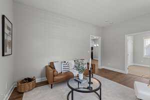 Living room with a brown leather sofa, striped pillow, and white throw. A round coffee table with books and candleholders sits on a light rug. A basket with blankets is beside the sofa. Doorways lead to other rooms, and a framed abstract art piece is on the wall.