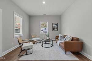 A living room with a brown leather sofa, two white upholstered chairs, a round glass coffee table, and a light gray area rug. Three decorative cushions rest on the sofa. Two windows and a framed picture are on the walls.