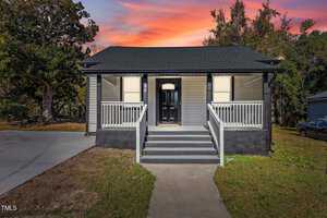 The single-story house at 703 Vance Street in Henderson features gray siding and black roofing. A charming front porch with white railings and a black door adds to its appeal. Surrounded by trees, the home is beautifully set against a sunset sky painted with vibrant pink and orange hues.