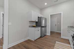 A modern kitchen with light gray walls, white cabinets, stainless steel refrigerator, granite countertops, a fruit bowl, and wood flooring. There is a door and an open doorway visible in the background.