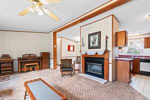 A living room with beige carpet and white walls features a ceiling fan, a piano, a fireplace with a painting above it, and a small armchair. The room is adjacent to a kitchen area with wooden cabinetry.