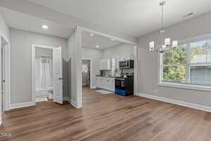 Open concept kitchen and dining area with light grey walls and wood flooring. The kitchen features white cabinets, stainless steel appliances, and a window. Adjacent is a small bathroom with a shower. A chandelier hangs above the dining space.