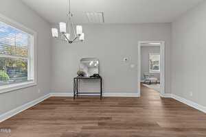 An empty room with light gray walls and wood flooring. A six-light chandelier hangs from the ceiling. A small table with a mirror and a decorative object is against the wall. A doorway leads to another room with a chair and window.