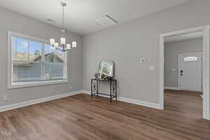 A room with light gray walls and wood flooring, featuring a window with blinds, a ceiling light fixture, and a small table with decor items beneath a wall-mounted mirror. An open doorway leads to another area with a white door.