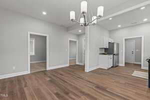 Empty room with hardwood floors, beige walls, and recessed lighting. A chandelier hangs from the ceiling. A small kitchen area with white cabinets and a gray countertop is visible, along with several doorways leading to other rooms.