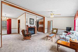 A spacious living room with carpeted floors features a fireplace with a wooden mantel, an armchair, a sofa, and a rocking chair. A ceiling fan is mounted above. The room leads to a dining area and a kitchenette. Red curtains adorn the windows.