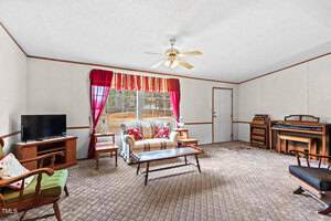 A living room with a patterned carpet features a sofa, armchairs, and a wooden coffee table. A ceiling fan hangs above. A TV sits on a stand, and a musical organ is in the corner. Red curtains frame a large window letting in natural light.