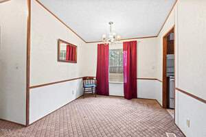 A small dining room with beige walls and a carpeted floor. A window with red curtains is centered on one wall, with a wooden chair placed beneath it. A chandelier hangs from the ceiling, and a rectangular mirror is on the side wall.