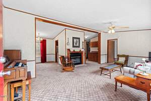 Living room interior with beige carpet and walls. A ceiling fan is above, and there's a fireplace with a wooden mantle. Furniture includes a mix of chairs, tables, and a couch. An open doorway leads to another room, and a piano is visible on the left.