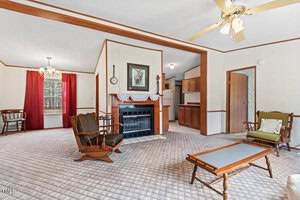 A living room with carpeted flooring, a ceiling fan, and a fireplace with a framed picture above. Wooden furniture includes rocking chairs and a coffee table. There's a view into a kitchen and dining area with red curtains and a chandelier.