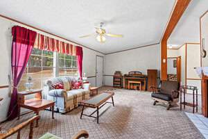 A living room with patterned carpet, a white sofa with colorful cushions, two wooden chairs, and a glass coffee table. A ceiling fan is above, and red curtains frame the window. An organ and wooden furniture are against the wall.