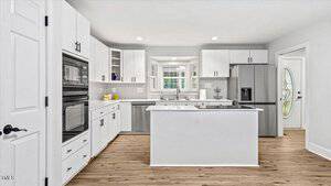A modern kitchen with white cabinets, a center island, and stainless steel appliances. A window is above the sink, and there is a door with an oval glass design on the right. The floor is wooden, and the walls are light-colored.