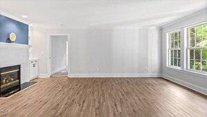 A spacious living room with light-colored walls and wood flooring. There is a large window on the right, providing natural light. A white fireplace with a blue accent wall and a decorative plate is on the left. A doorway leads to an adjacent room.
