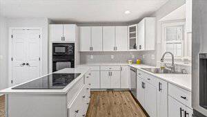 Modern kitchen with white cabinets and light wood flooring. Features a central island with a black cooktop. Stainless steel fixtures include a sink under a window. Double oven is built into the cabinets. Walls are light gray with soft lighting.