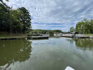 A calm lake scene with several docks and boats moored on either side. The foreground shows still water under a partly cloudy sky. Surrounding the lake are lush green trees and a few structures that seem like boathouses or sheds.