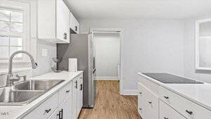 Modern kitchen with white cabinets and countertops. A stainless steel sink is on the left, next to a window. The kitchen features a large stainless steel refrigerator and an island with an electric cooktop. The floor is light wood.