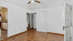 An empty room with white walls and ceiling. A ceiling fan with light is in the center. The floor is wooden. There is a white door with a frosted glass window propped open, leading into the space with more visible rooms beyond.