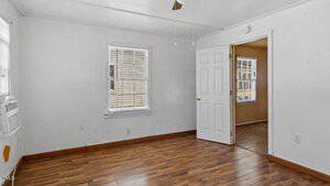 An empty room with two windows, wood laminate flooring, and a white ceiling fan. The walls are white, and there is a white door partially open, revealing a glimpse of an adjoining room with similar flooring. A wall-mounted air conditioner is visible.