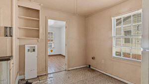 Empty room with beige walls and tiled floor featuring a built-in shelf and a window on the right. A doorway leads to another room with wooden flooring and a single window, visible in the background. Ceiling fan parts are visible at the top.