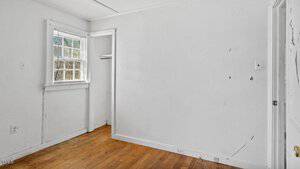 An empty room with white walls and a wooden floor. There is a window on the left with blinds partially open, a small open closet, and a white door on the right. The walls show signs of minor wear and scuff marks.