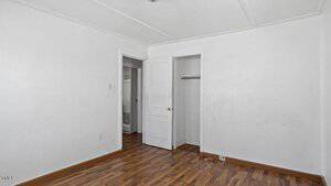 An empty room with white walls, a wooden floor, and a closed white door. There is an open closet with no door on the right and a slightly open door leading to another room on the left. The ceiling has a light fixture.