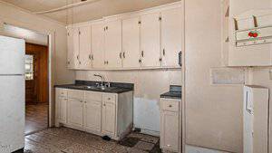 A small, beige kitchen with outdated cabinets and a black countertop. There's a stainless steel sink, missing lower cabinet panels, and a broken floor tile in front. A white refrigerator is partially visible on the left.