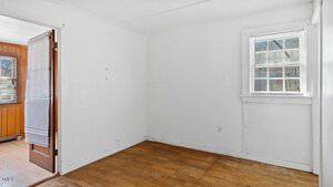 Small empty room with white walls and a wooden floor. There is one window with blinds partly open, allowing natural light. An open door with blinds leads to another area with wooden paneling visible.
