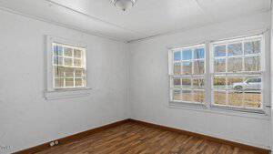 An empty room with white walls and a wooden floor. There are two windows, one on each wall, allowing natural light into the space. A light fixture is mounted on the ceiling. A car is visible outside through the right window.
