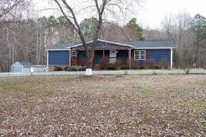 A single-story blue house with a wooden porch is situated in a wooded area. The yard is covered with fallen leaves, and a bare tree stands in front. A small shed is visible to the left, and the background features trees with sparse foliage.