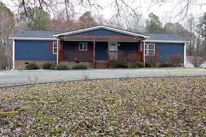A blue ranch-style house with a brick foundation and white trim. It has a porch with wooden railings and a gravel driveway in front. The yard is bordered by trees, and the ground is covered with dry leaves.