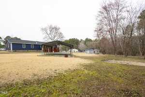 A yard with a metal carport beside a blue house. The ground is covered in brown grass and patches of soil. Leafless trees surround the area, and a few sheds are visible in the background. The sky is overcast.