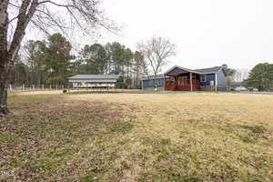 A blue house with a wooden porch stands on a spacious lawn with sparse grass. There is a fenced area and a shed to the left, surrounded by trees. The sky is overcast.