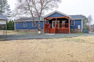 A blue single-story house with a wooden porch and steps is centered in the image. The yard in the foreground has brown grass, and a large tree stands nearby. The porch has a railing and is elevated. The sky is overcast.
