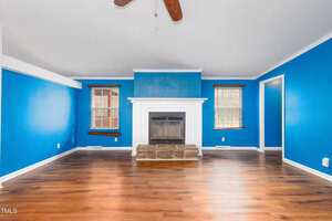 A living room with bright blue walls, a white fireplace centered on one wall, and two windows with wooden blinds. The floor is wood with a natural finish. A ceiling fan with wooden blades is visible above.