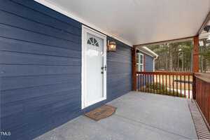 A blue house exterior features a white door with an arched window. A hanging lantern light is on the wall next to the door. The wooden porch has a mat labeled "HOME" and is enclosed by a wooden railing. Trees are visible in the background.