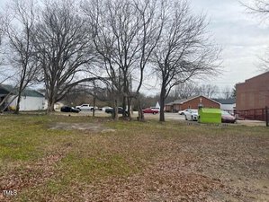 A grassy area with scattered fallen leaves is in the foreground at 313 Piedmont Avenue, Oxford. In the background, several bare trees stand beside a row of parked cars and trucks, along with a green dumpster and brick buildings. The sky remains overcast.