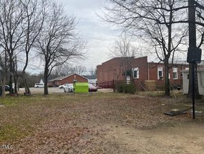 A view of a yard with bare trees and a basketball hoop on the right. In the background, there are red brick buildings, a lime green dumpster, and several parked vehicles, including trucks and cars, near a driveway.