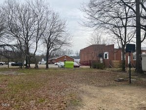 A vacant lot with sparse grass and trees in the foreground. A green dumpster is visible near red brick buildings in the background. Several parked vehicles are on the left. The sky is overcast.