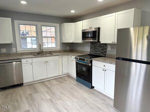 Modern kitchen with white cabinets, granite countertops, and a gray tile backsplash. Stainless steel appliances include a dishwasher, oven, microwave, and refrigerator. Two windows provide natural light. Light wood flooring completes the look.