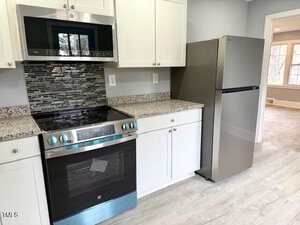 A modern kitchen featuring a stainless steel refrigerator, oven, and microwave. White cabinets and a speckled gray countertop are present. A black and gray mosaic tile backsplash is above the stove. A windowed room is visible in the background.