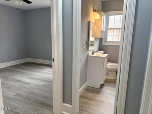 View of a small bathroom with a white vanity and toilet, situated beside an empty room with light gray walls and wooden flooring. The bathroom has a window above the toilet and a mirror above the vanity, with a ceiling fan visible in the adjoining room.