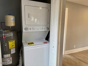 Stacked white washer and dryer unit in a closet with an open door. A water heater is located to the left with visible pipes. The floor has light wood flooring, and the walls are painted gray.