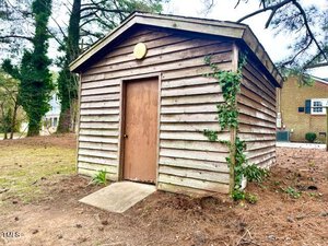 A small, weathered wooden shed stands on a grassy area surrounded by trees. The shed has a brown door and a shingled roof. Ivy is growing on one side of the shed. A concrete step leads to the entrance. A brick building is partially visible in the background.