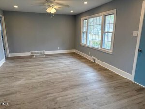 A spacious, empty room with light wood flooring and gray walls. The room features a ceiling fan with lights, a blue door, and large windows allowing natural light. White trim accents the windows and baseboards. A vent is located on one wall.