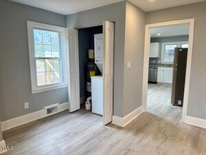 A room with light gray walls and wood flooring. A small laundry area with a washing machine and dryer is behind folding doors. Two windows and a door lead to a kitchen with white cabinets and a dark fridge. Natural light illuminates the space.