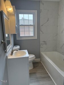 A small bathroom with light blue walls and a window. It features a white sink with a cabinet and mirror above, a toilet, and a bathtub with a light gray and white marble-patterned wall. The floor is gray, and a wall-mounted light is above the mirror.