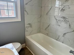 A bathroom with a white bathtub against a wall of gray and white marble-patterned tiles. A white toilet is adjacent to the tub. A window with white trim is on the left, allowing natural light into the room.