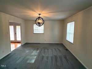 An empty room with light-colored walls and carpeted floor. A decorative ceiling light fixture is centered. Two windows allow natural light. A doorway opens to another room with a hardwood floor.