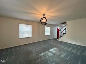 A spacious living room with light gray carpet and white walls. Two windows provide natural light. A black chandelier hangs from the ceiling. A staircase with black railing leads upstairs, and a red door is visible in the background.