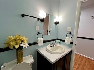 Bathroom with a simple sink and counter setup. Two wall lights flank a rectangular mirror above the sink. Towel holders with towels are on both sides. A vase with gold flowers sits on the toilet tank. Wood flooring extends into the hallway.